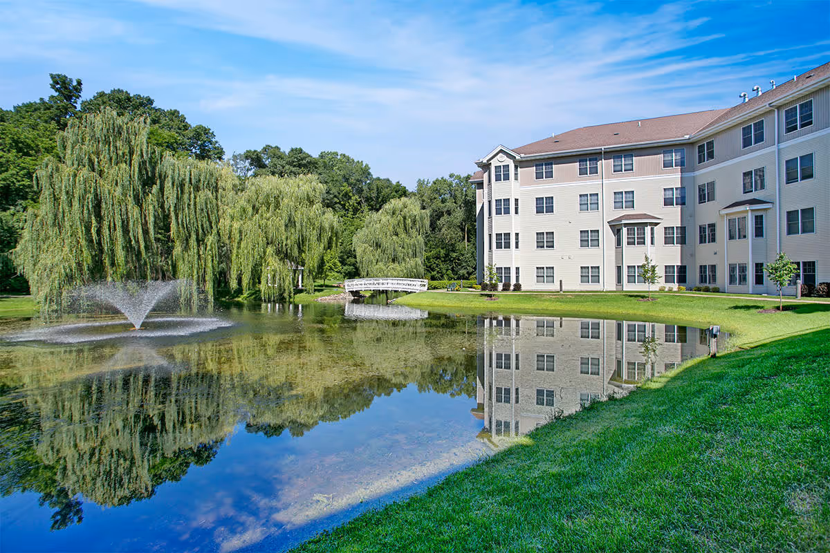 A multi-story senior living building beside a pond with a fountain, a small white footbridge, and willow trees reflected in the water.