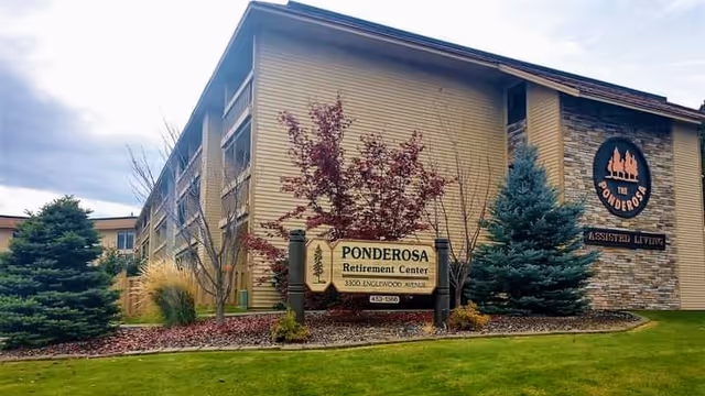 Exterior view of Ponderosa Retirement Center building with a sign in front displaying the name and address. The building has beige siding with stone accents and several trees and shrubs in the landscaped area around it.