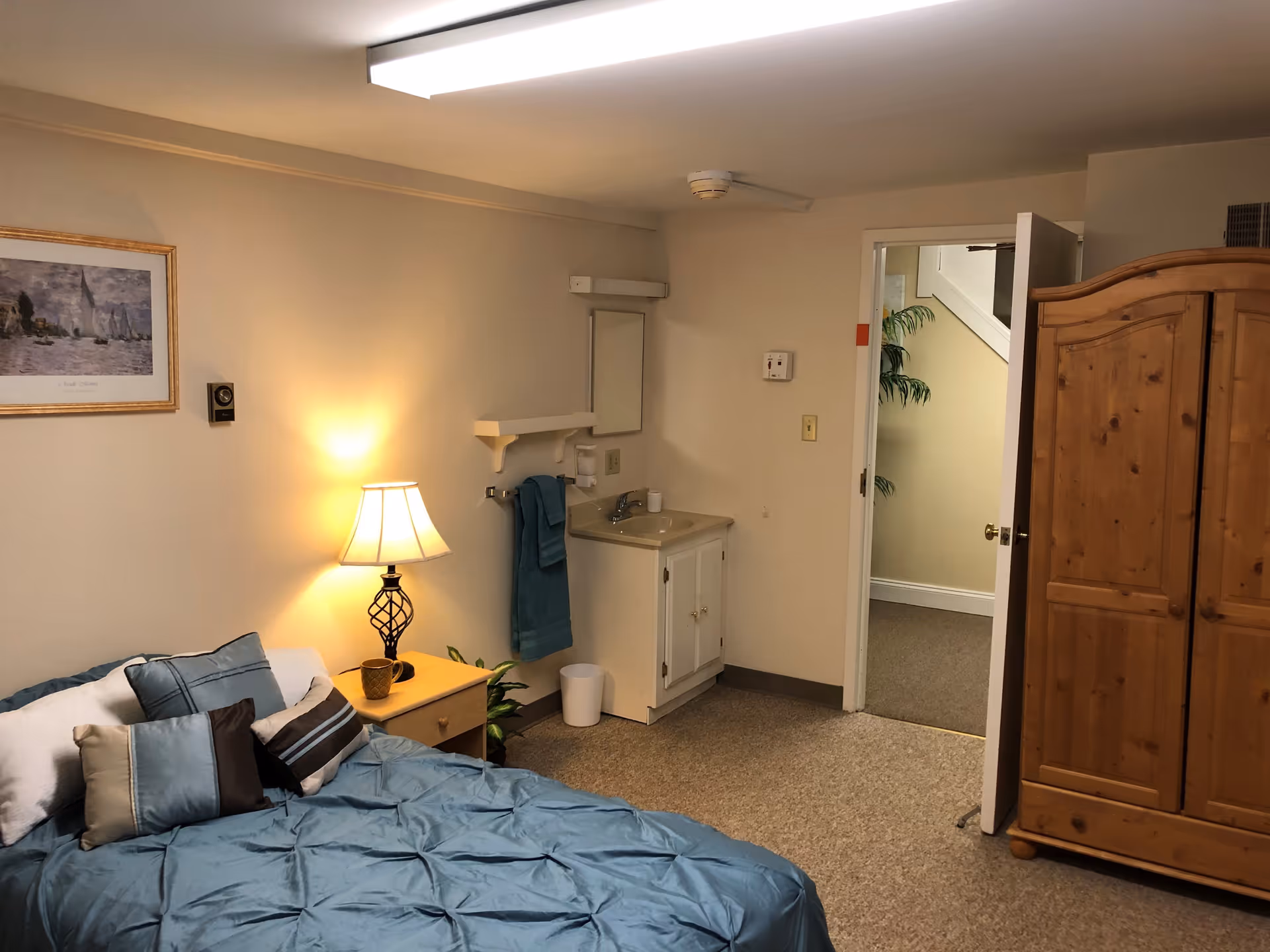 A cozy bedroom in The Heritage of Green Hills facility featuring a bed with blue and brown pillows and a blue comforter. Next to the bed is a wooden nightstand with a lamp and a small plant. On the wall above the bed is a framed picture. There is a small sink with a mirror and towel rack on the adjacent wall. A wooden wardrobe stands near an open door leading to a hallway with a plant visible.