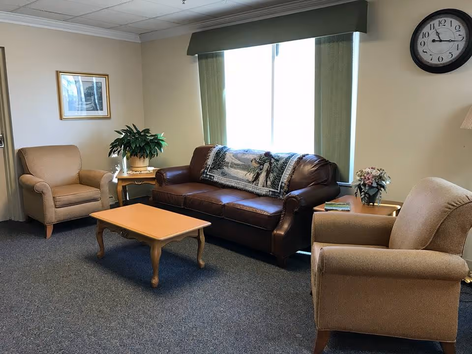 A cozy living room area with a brown leather sofa adorned with a decorative throw, two beige armchairs, a wooden coffee table, and side tables with plants and flowers. A large window with green curtains lets in natural light, and a clock is mounted on the wall above one armchair.
