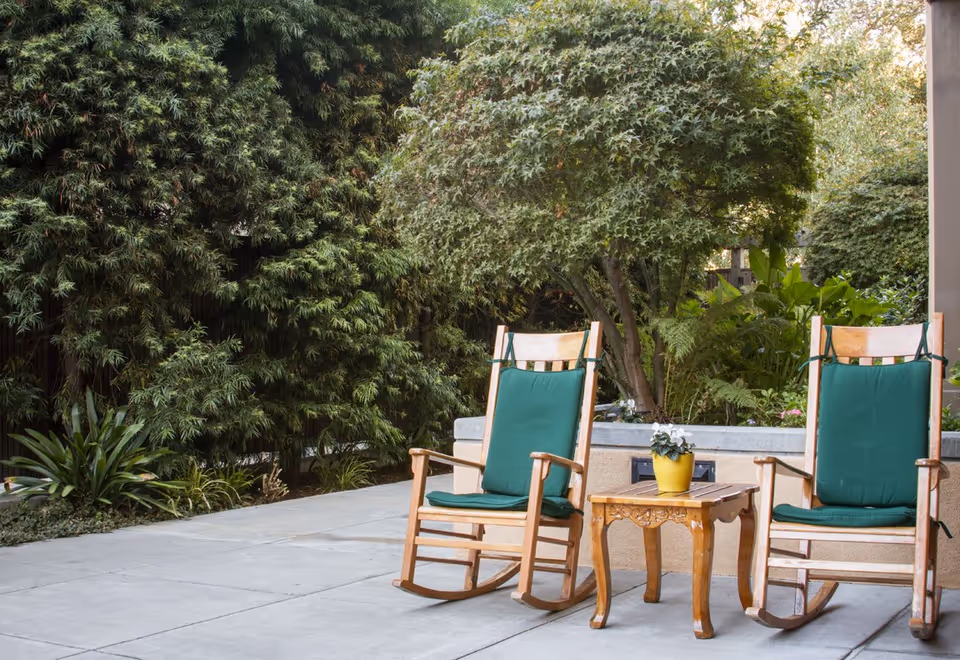 Two wooden rocking chairs with green cushions on a concrete patio next to a small wooden table with a yellow pot containing a white flower, surrounded by lush green trees and plants.