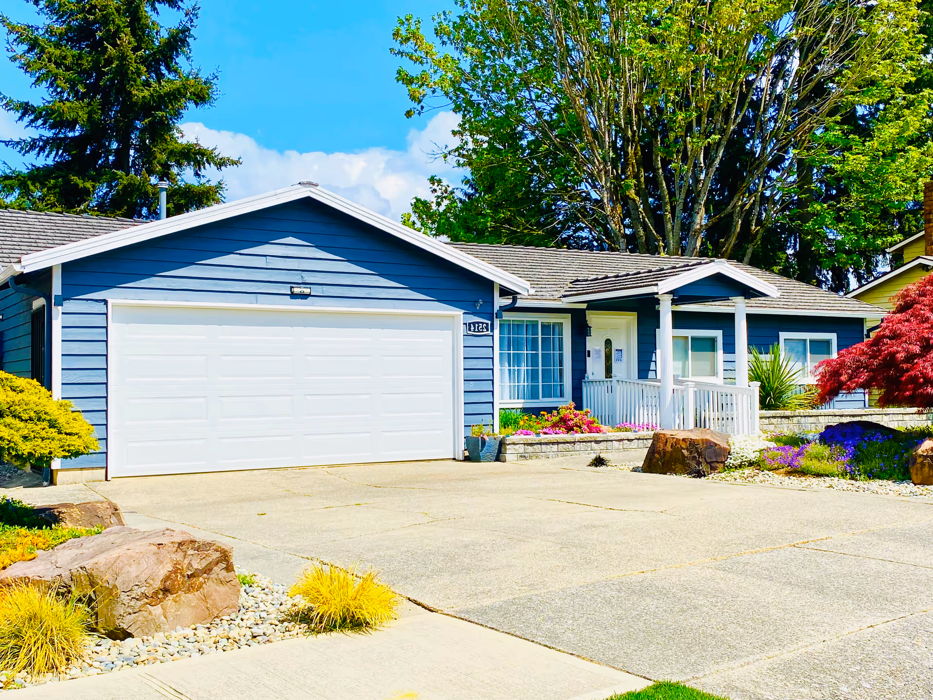 Front exterior of a single-story blue house with a two-car garage, a small porch, and landscaped yard.