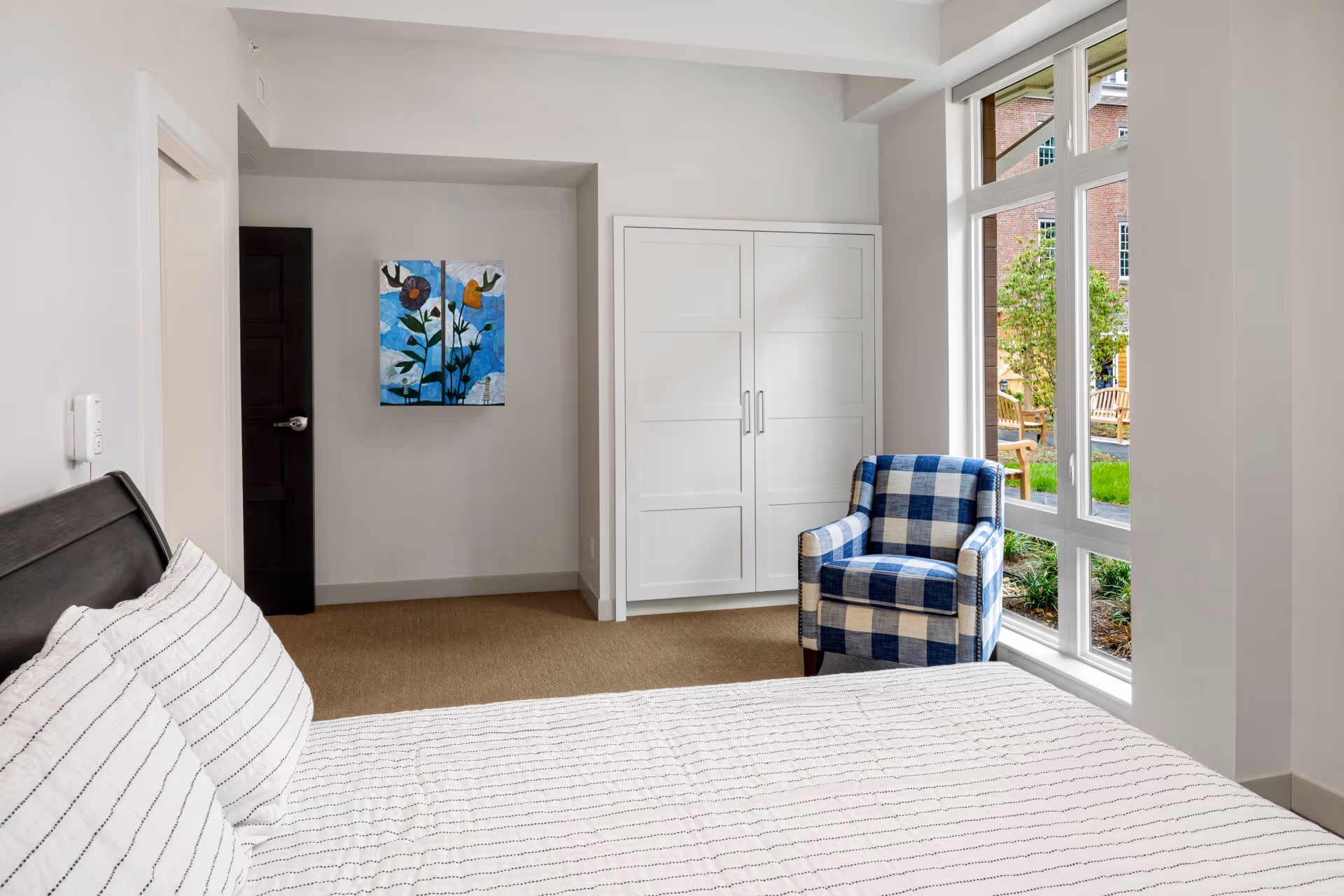 A bright bedroom with a bed featuring white and black striped bedding, a blue and white checkered armchair near a large window overlooking a garden with benches, a white wardrobe, and a colorful floral painting on the wall next to a dark wooden door.