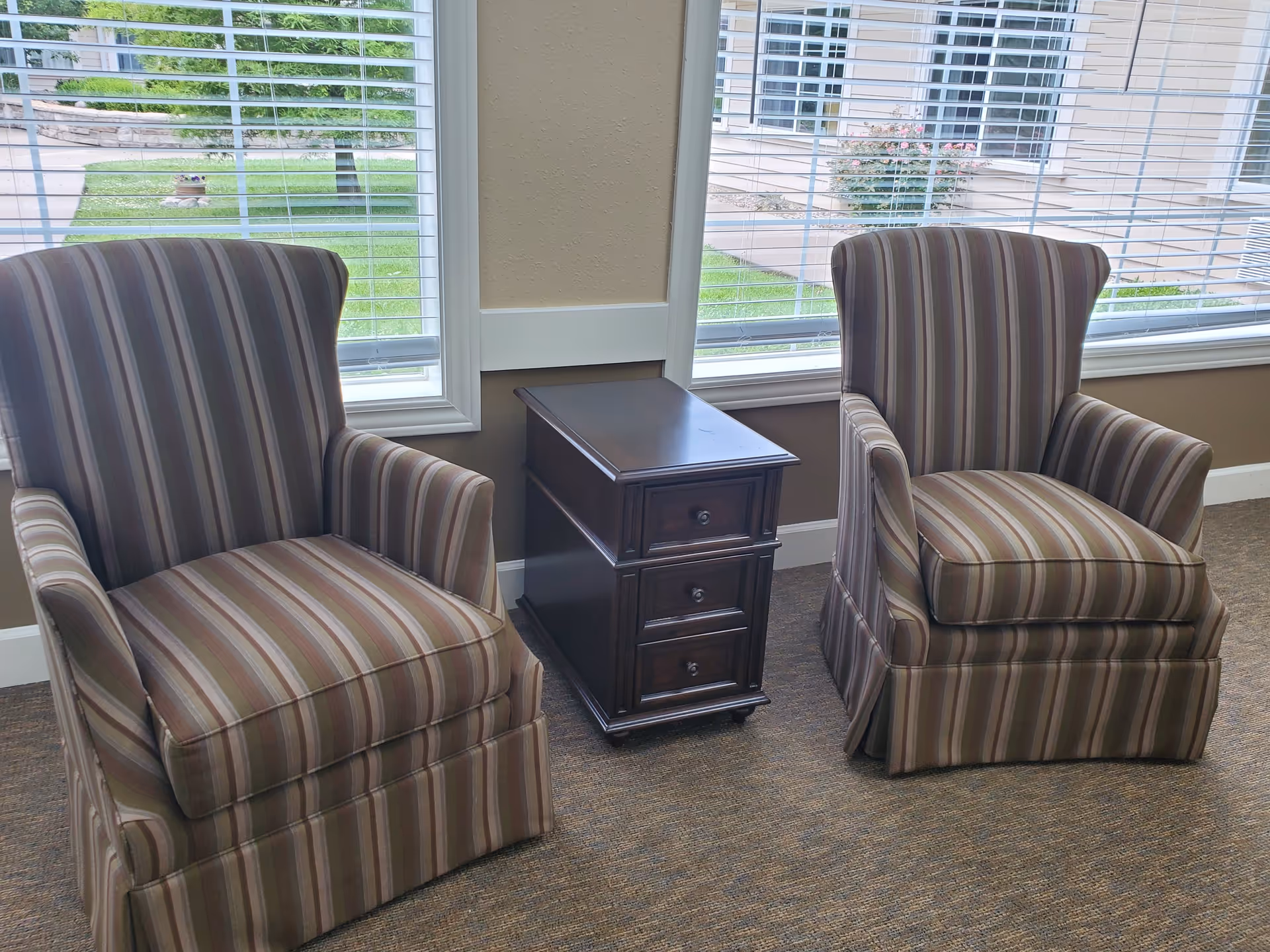 Two striped upholstered armchairs with a small dark wooden side table with three drawers between them, placed in front of large windows with white blinds. Outside the windows, green grass and parts of a building are visible.