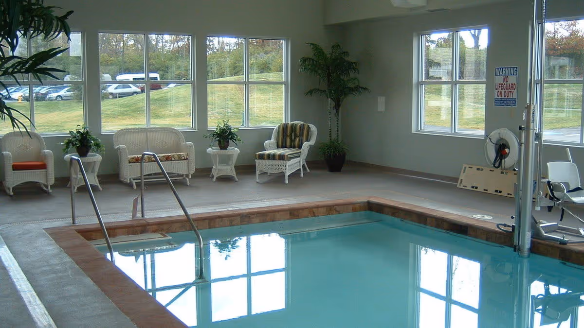 Indoor swimming pool area with clear water, metal handrails, and seating including wicker chairs and small tables with plants. Large windows provide a view of the grassy outdoor area and parked cars. A sign on the wall reads 'Warning No Lifeguard On Duty'.