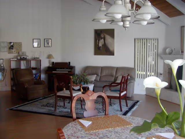 A living room area with a gray sofa, wooden chairs, a small table with a plant, a brown armchair, a rug, and a painting of a horse on the wall. In the foreground, there is a round table with a floral tablecloth and a vase with white calla lilies. A chandelier with multiple lights hangs from the ceiling.