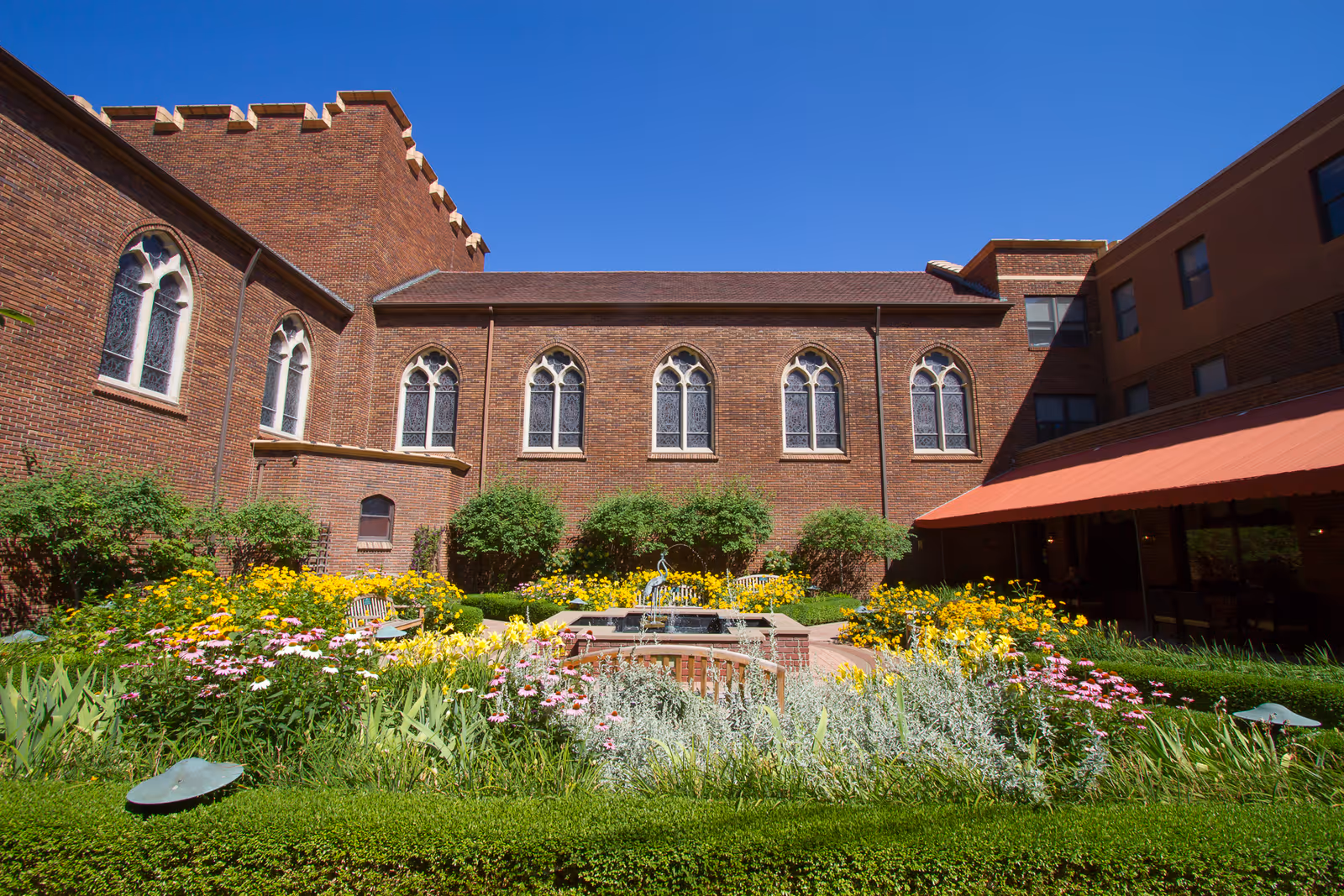 Sunlit courtyard garden with a central fountain surrounded by colorful flowers and hedges, bordered by a brick building with arched windows under a clear blue sky.