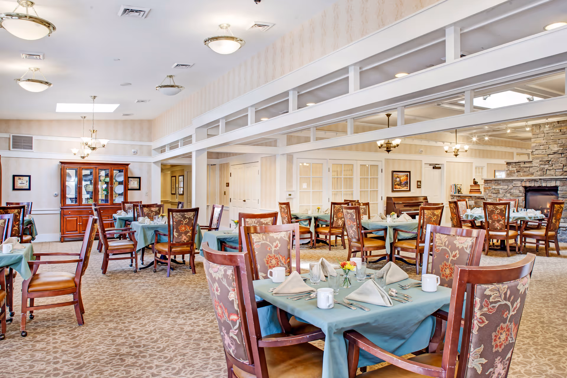 Spacious dining room in a senior living facility with tables set for meals, upholstered wooden chairs, and a stone fireplace.