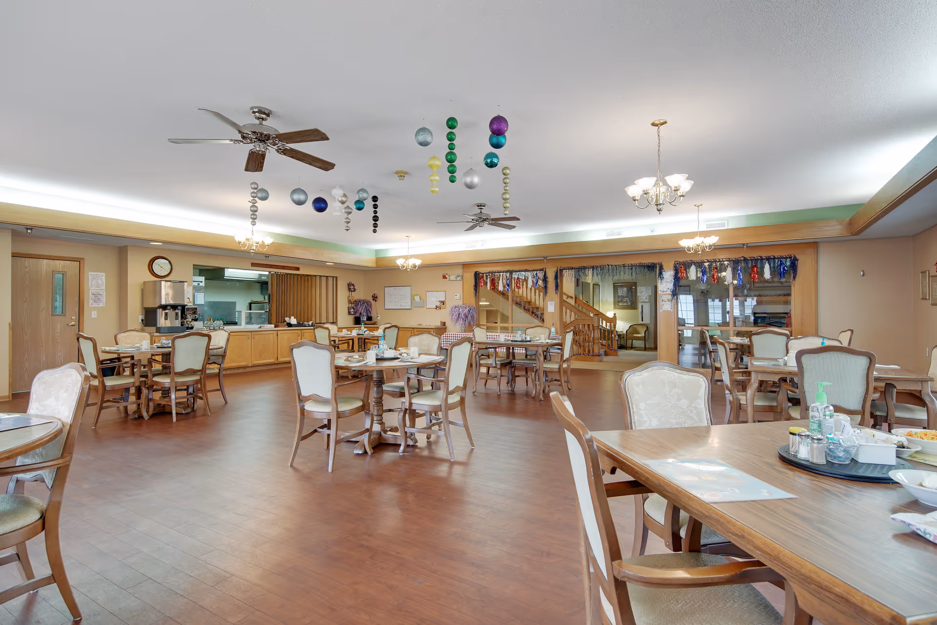 A spacious dining room in Elmhurst Commons with multiple wooden tables and cushioned chairs arranged neatly. The room has a wooden floor, ceiling fans, and decorative hanging ornaments from the ceiling. There is a serving counter area with a coffee machine and kitchen visible in the background. The room is well-lit with chandeliers and natural light coming through windows.