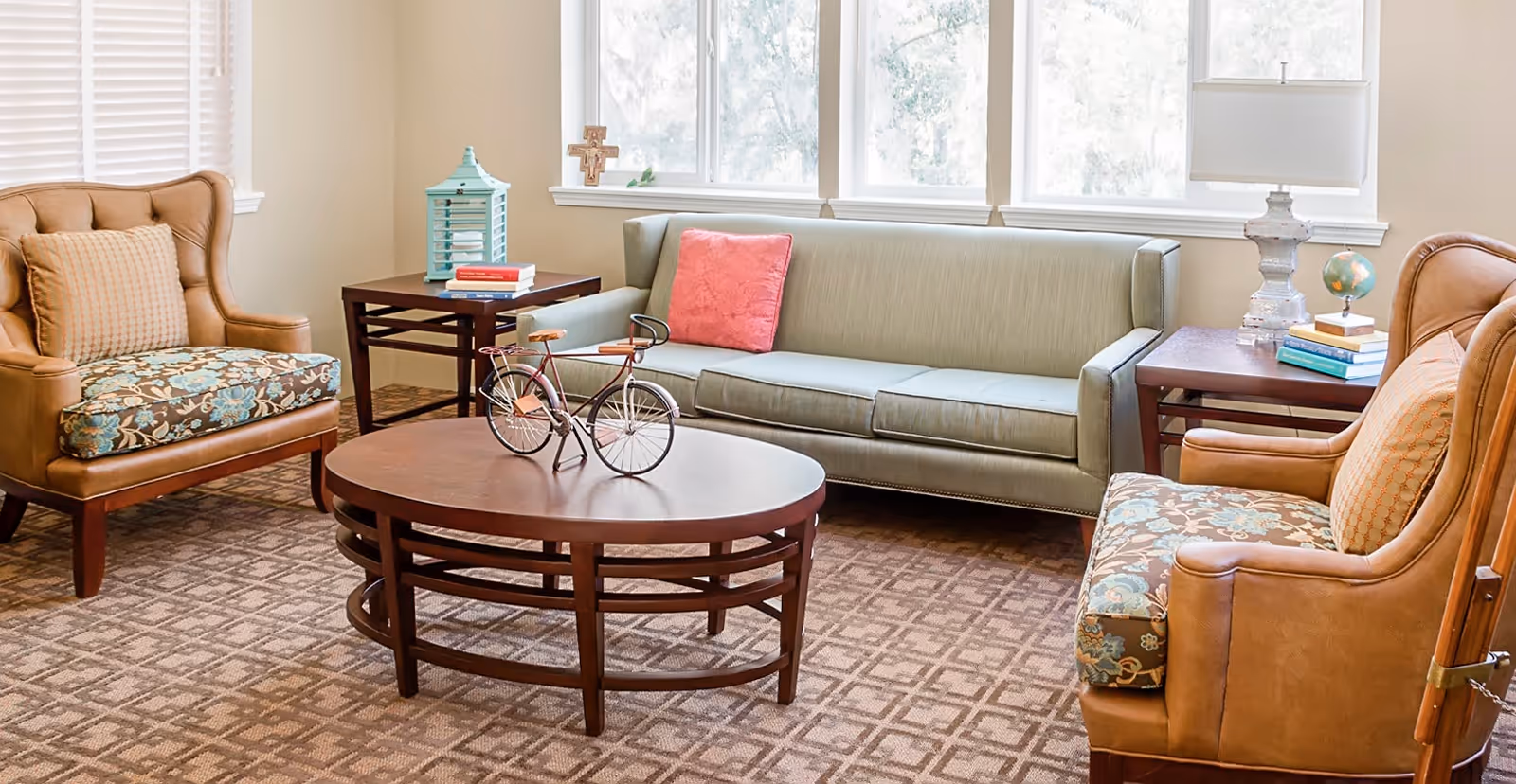 A bright living room with a green sofa, two upholstered armchairs, a round wooden coffee table and side tables in front of large windows.