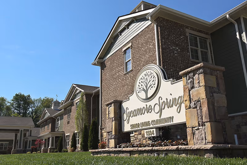Exterior view of Sycamore Springs Senior Living Community building with a large sign in front displaying the community name and address. The building has brick and siding walls, multiple windows, and a clear blue sky above.