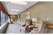 Sunlit interior corridor with upholstered chairs, a small side table and lamp along a row of windows.