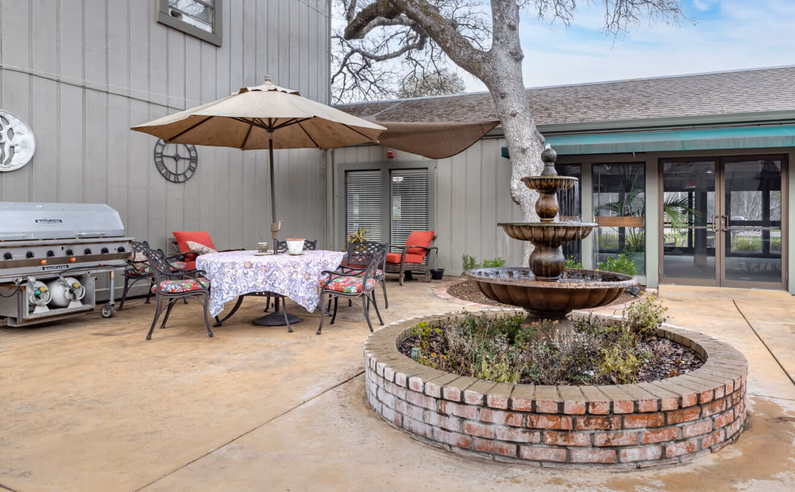 Brick circular fountain in a courtyard with a patio table and umbrella, chairs, and a grill near the building entrance.
