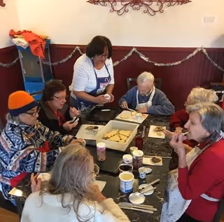 A group of elderly people sitting around a table in a room, engaging in a cookie decorating activity with an instructor standing and assisting them. The table is covered with various decorating supplies and cookies.
