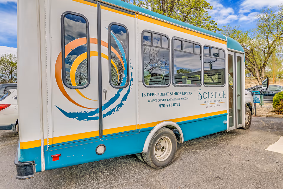 Side view of a white and teal shuttle bus with yellow and orange circular designs, parked outdoors near trees and other vehicles. The bus has text advertising Solstice Senior Living at Mesa View, including their website and phone number.