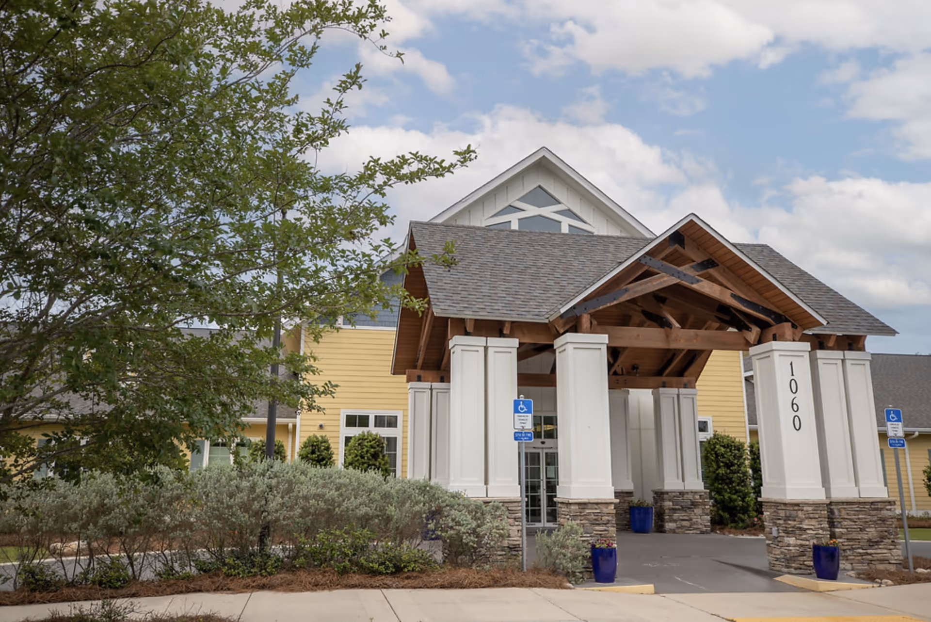 Exterior view of a senior living facility entrance with a covered driveway supported by white pillars with stone bases. The building has yellow siding and a gray shingled roof. There are blue handicap parking signs and blue flower pots near the entrance. Trees and shrubs surround the area under a partly cloudy sky.