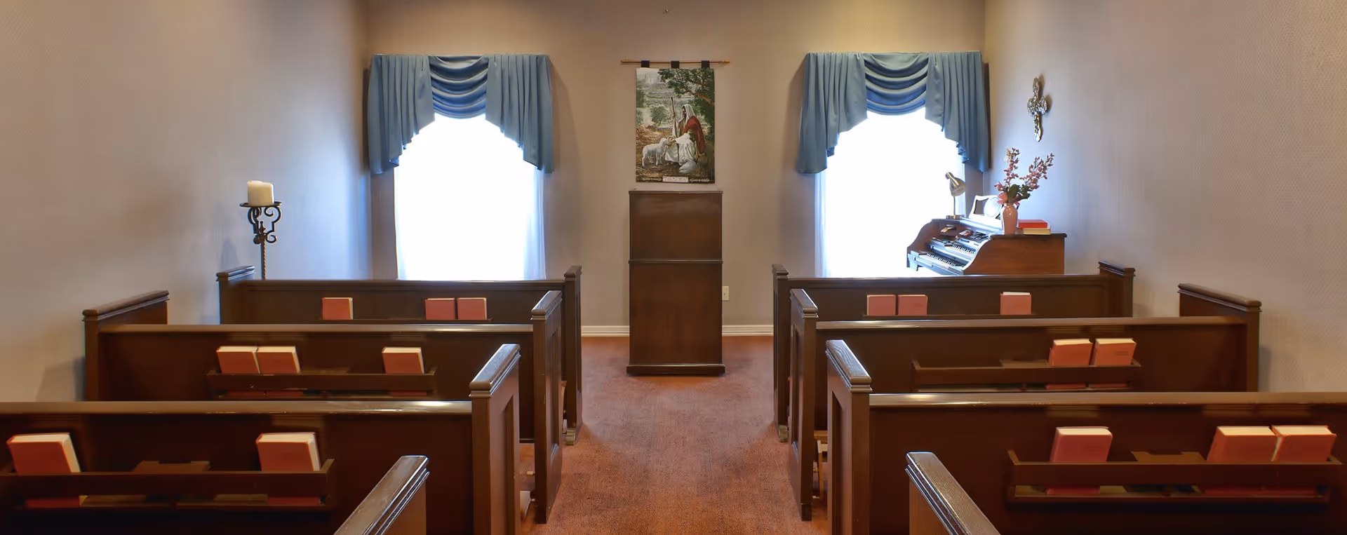 Small chapel room with wooden pews arranged in two rows facing a wooden podium. Two windows with blue valance curtains are on the back wall, one on each side of the podium. A religious tapestry hangs above the podium, and a small organ with flowers and books is positioned near the right window. A candle holder with a lit candle is on the left side of the room.