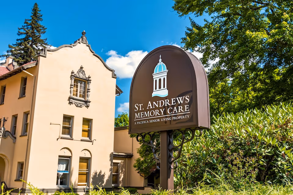 Outdoor view of St. Andrews Memory Care facility sign with a beige building and green trees in the background under a blue sky with some clouds.