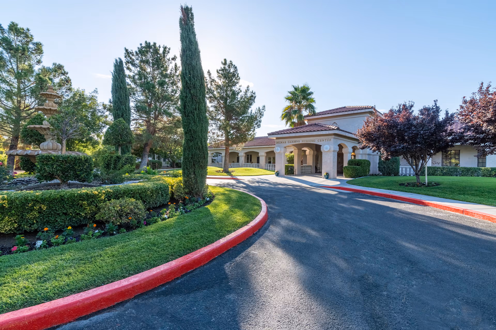 Exterior view of a nursing center building with a curved driveway bordered by red curbs. The building has a covered entrance with columns and a sign that reads 'Delmar Gardens Nursing Center'. The surrounding area features well-maintained landscaping with trimmed bushes, tall trees, a fountain, and green grass under a clear blue sky.