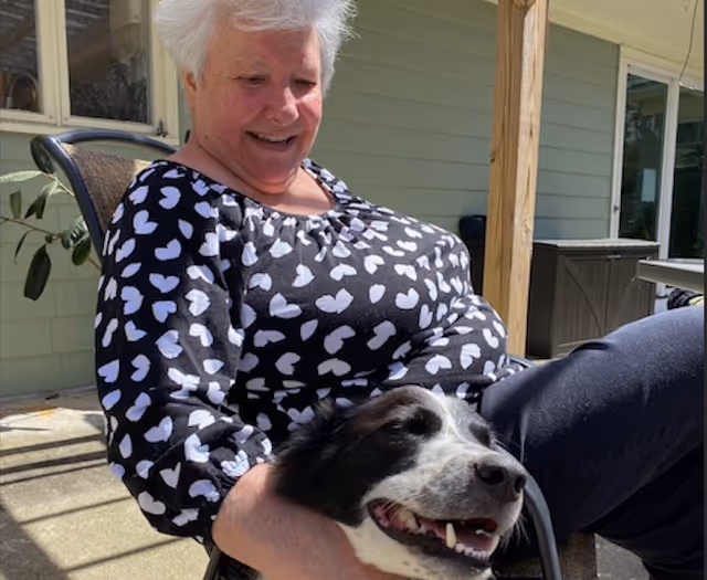 An elderly woman with short white hair wearing a black shirt with white heart patterns is sitting outdoors on a chair, smiling and holding a happy black and white dog on her lap. The background shows a light green building exterior with windows and a wooden post.