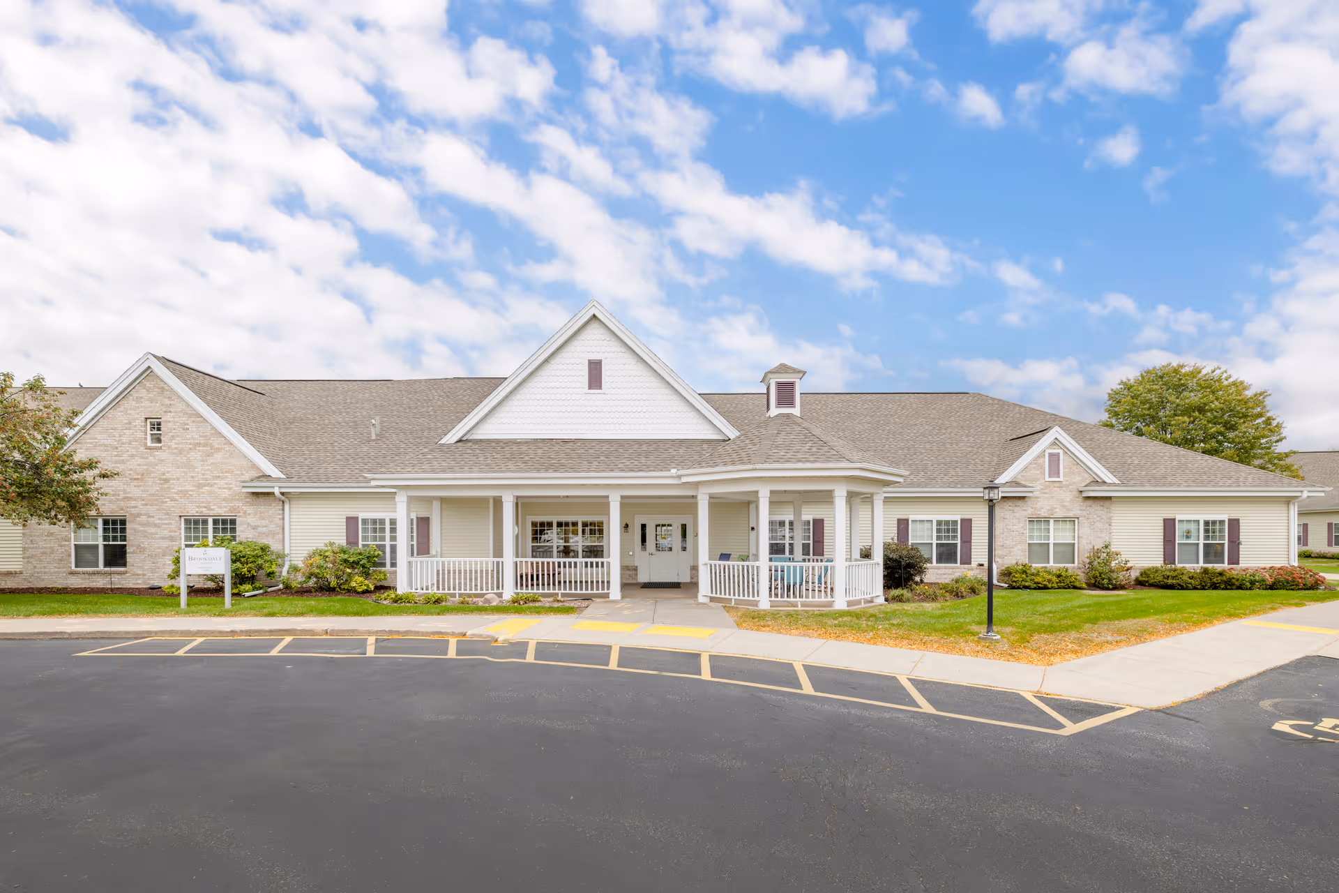 Exterior view of a single-story senior living facility building with a covered entrance porch, surrounded by a well-maintained lawn and a paved parking area under a partly cloudy sky.