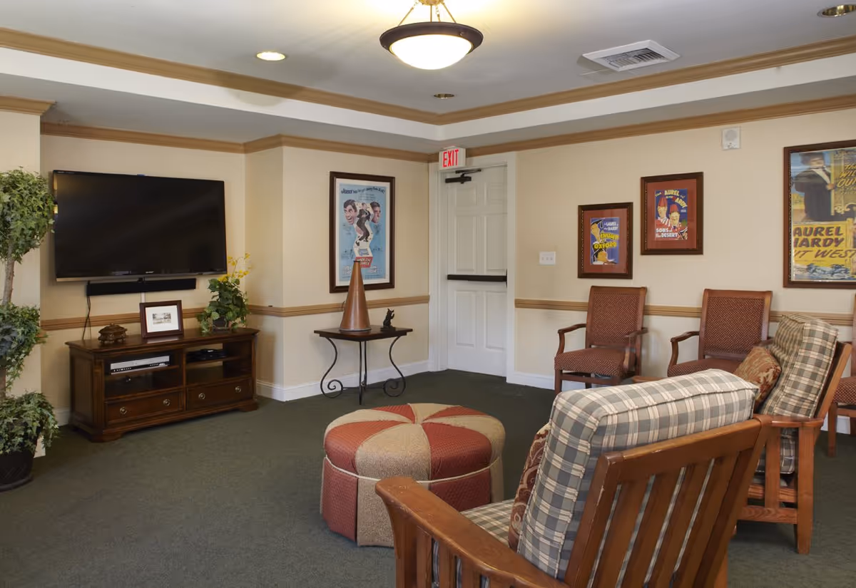 A cozy living room area with a flat-screen TV mounted on the wall above a wooden media console. The room features a round ottoman, two plaid cushioned wooden chairs, and two additional wooden chairs with red upholstery. The walls are decorated with framed vintage posters, and there is a small table with decorative items in the corner. The room has beige walls with wooden trim and a green carpeted floor.