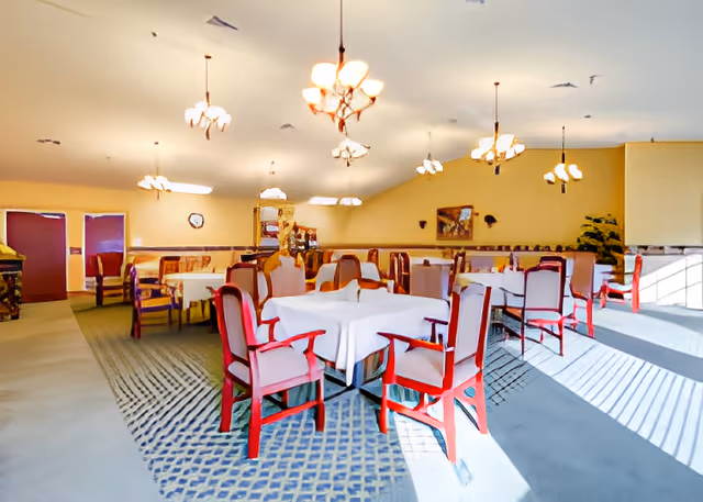 A spacious dining room with multiple tables covered in white tablecloths and surrounded by red wooden chairs with cushioned seats. The room is well-lit with several hanging chandeliers and natural light coming through large windows on the right side. The walls are painted a soft yellow, and there is a carpet with a patterned design on the floor. A clock and framed artwork are visible on the walls.