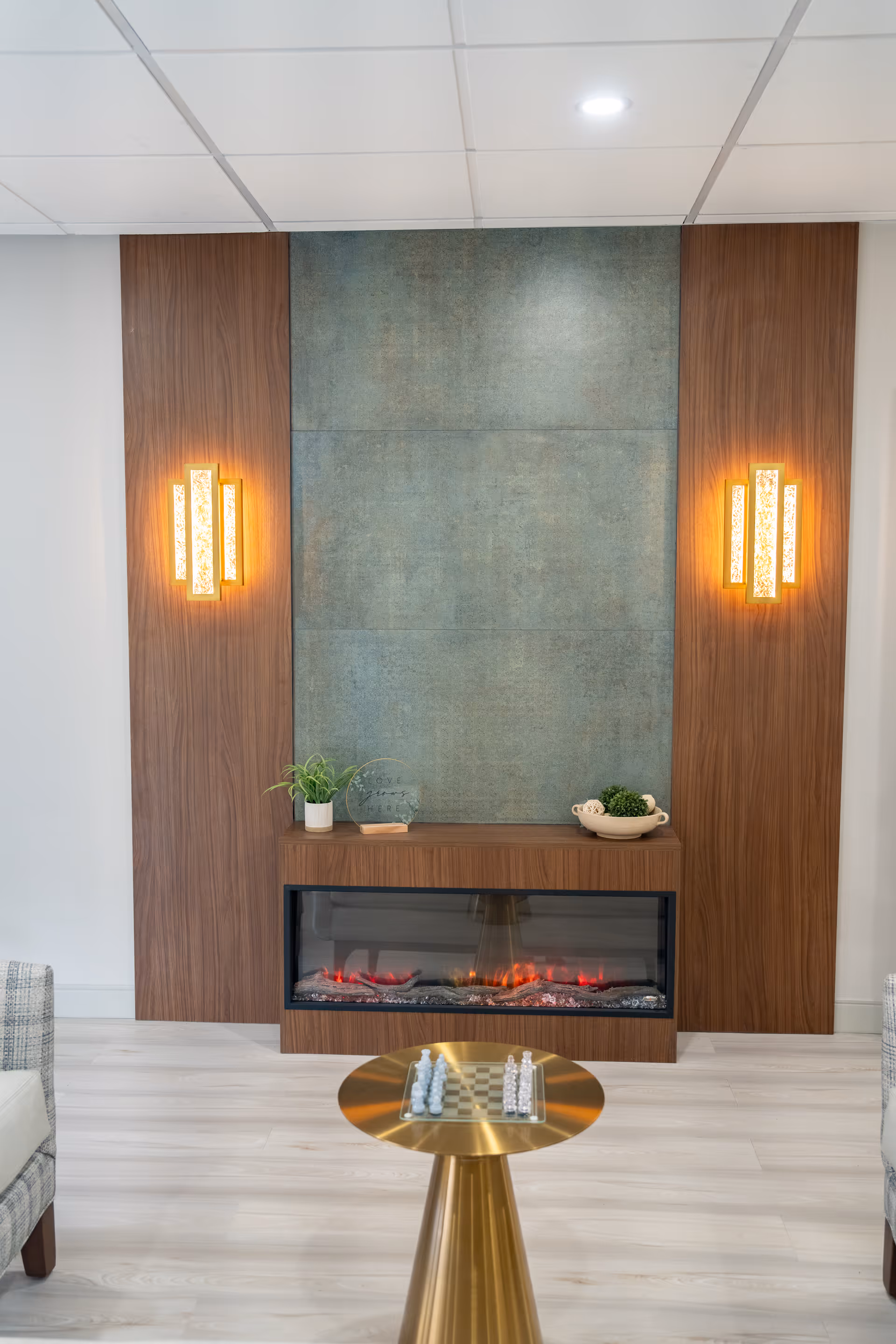 A modern indoor seating area featuring a built-in electric fireplace with a wooden and textured wall backdrop. Two vertical wall sconces with warm lighting flank the fireplace. A small round gold table with a chessboard and chess pieces is positioned in front of the fireplace. Part of two upholstered chairs are visible on either side of the image.