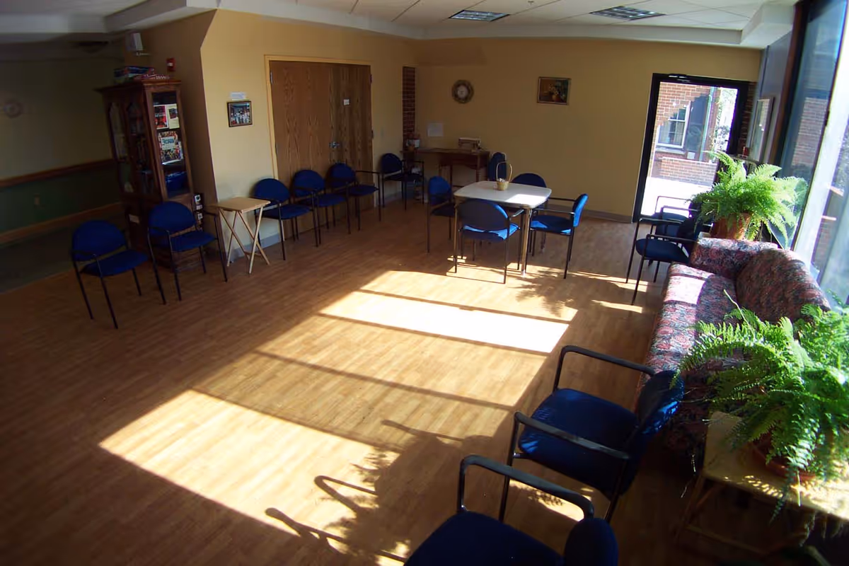 A well-lit common room with wooden flooring featuring a variety of seating options including blue chairs arranged along the walls and around a small square table. There is a floral-patterned couch near large windows letting in sunlight, and several potted ferns on tables. The room has light yellow walls with some framed pictures and a wooden cabinet in the corner.