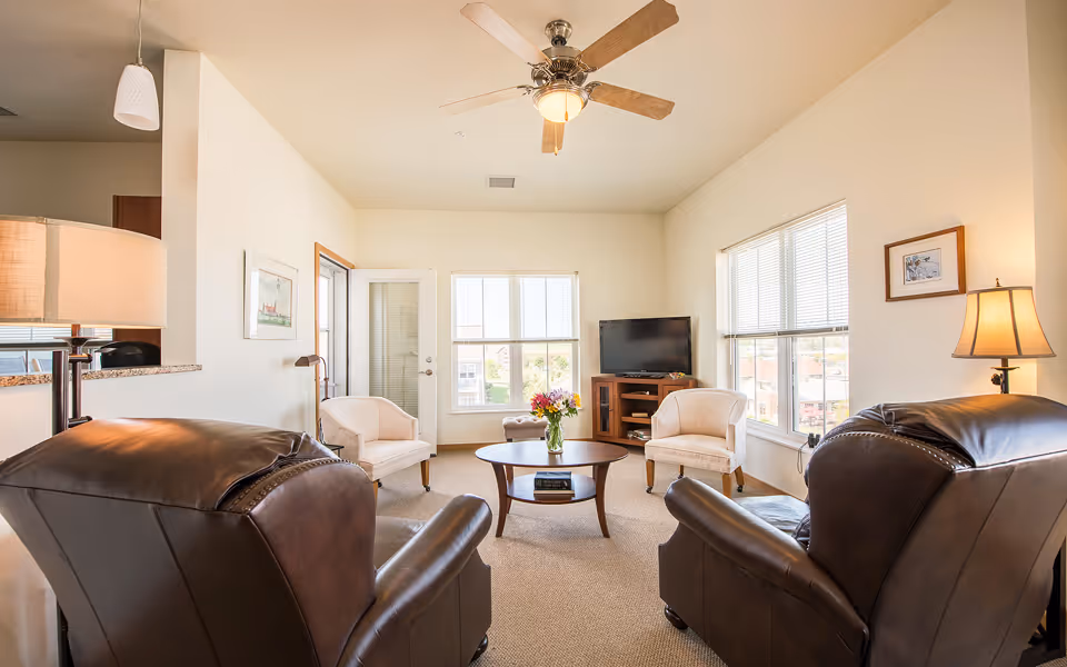 A bright and cozy living room with two large brown leather armchairs facing a round wooden coffee table with a vase of flowers. Two beige upholstered chairs are placed near large windows letting in natural light. A flat-screen TV sits on a wooden stand in the corner, and a ceiling fan with light is mounted above. The room has beige carpet and light-colored walls with framed artwork and a floor lamp.