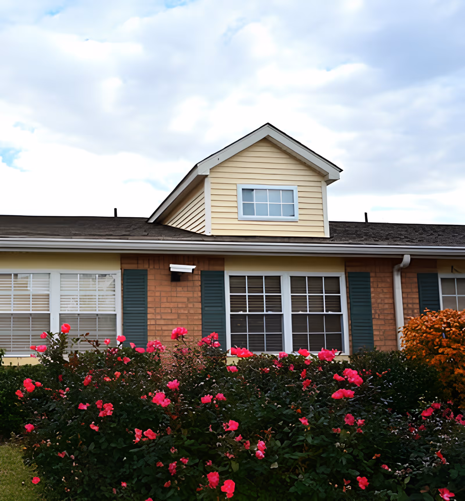 Front exterior of a one-story brick-and-siding building with windows, green shutters, and pink flowering bushes under a cloudy sky.