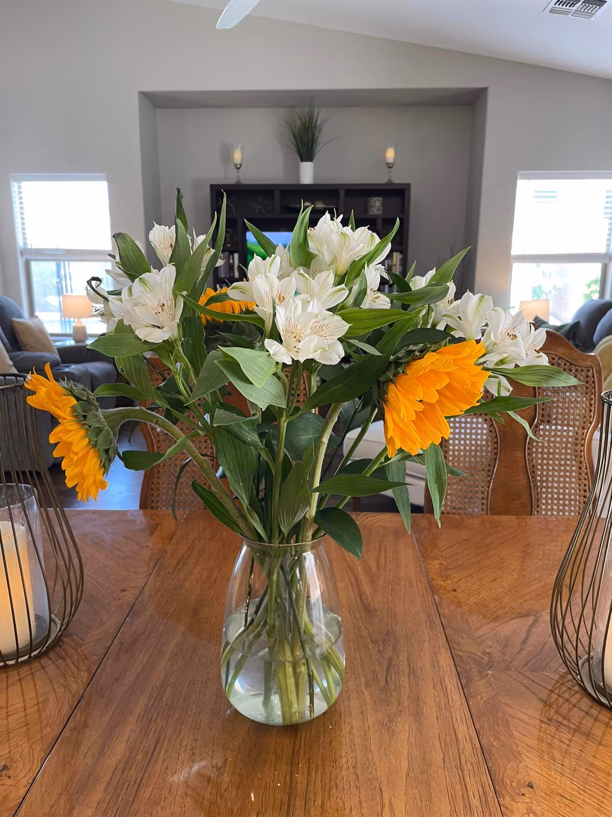 A clear glass vase with a bouquet of white and yellow flowers sits on a wooden dining table. The background shows a living room area with windows, chairs, a dark wooden shelving unit with decorative items, and two lit candles in wire candle holders on either side of the vase.