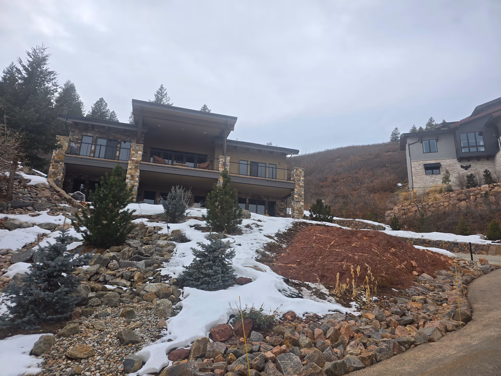 Exterior view of a multi-level building with stone pillars and balconies, surrounded by a rocky landscape with patches of snow and small evergreen trees under a cloudy sky.