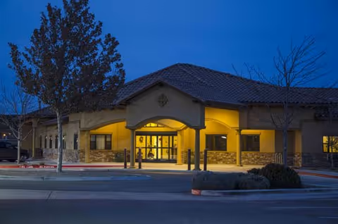 Front entrance of a single-story assisted living building illuminated at dusk with a driveway, trees, and a tiled roof.