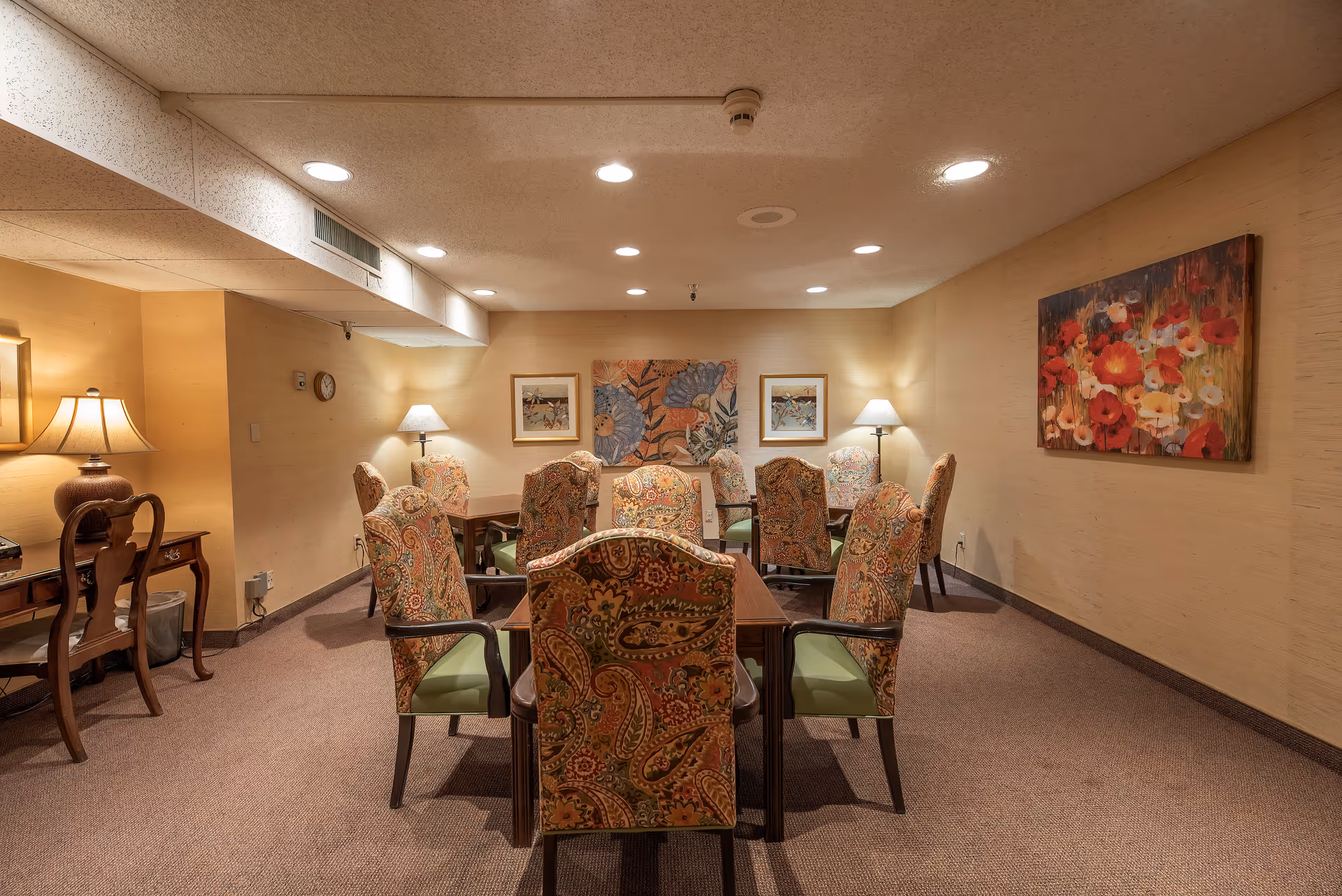 Carpeted communal dining room with a rectangular table surrounded by patterned upholstered chairs, warm lamps, and floral wall art.