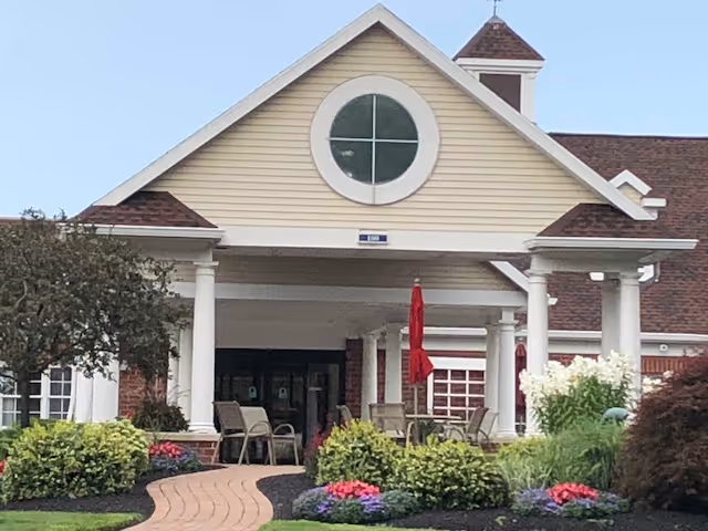 Front entrance of a senior living facility with a covered porch supported by white columns, a circular window above the entrance, outdoor seating with chairs and red umbrellas, and landscaped garden beds with colorful flowers and shrubs.