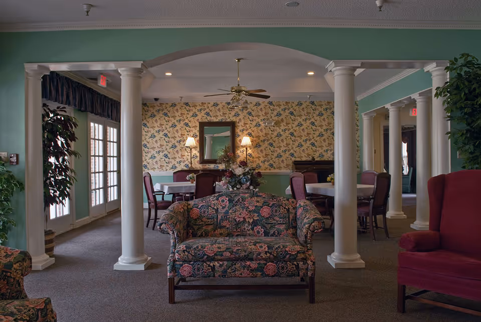 Interior view of a senior living facility lounge area with floral patterned loveseat in the foreground, white columns, green walls, floral wallpaper, ceiling fan, and tables with chairs in the background.