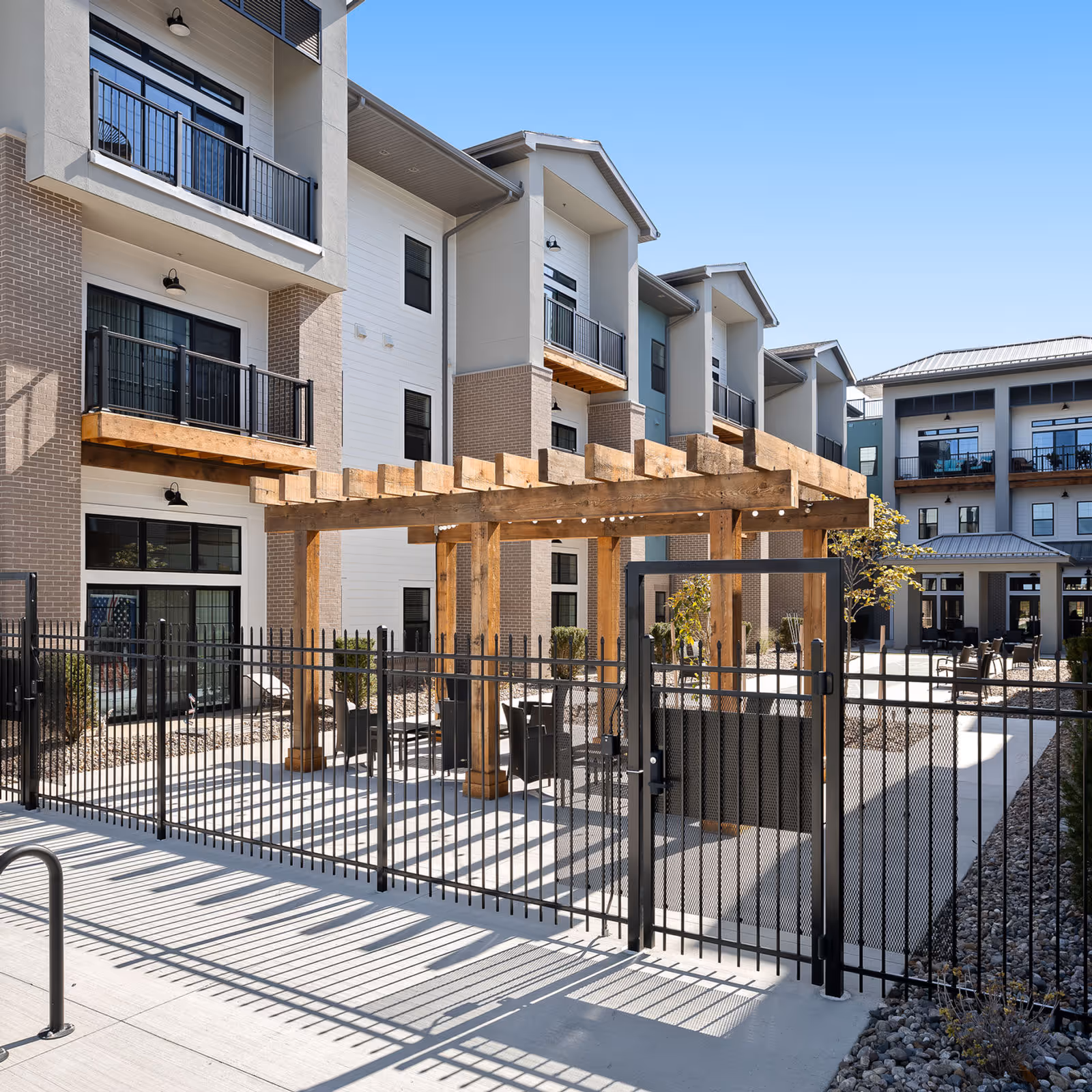 Courtyard of a modern senior living complex featuring a wooden pergola, gated fence, outdoor seating, and multi-story balconies.