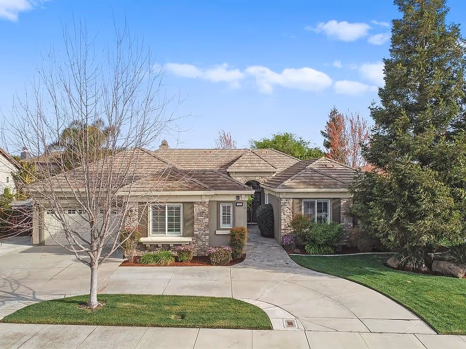 Front exterior view of a single-story residential building with a stone and stucco facade, a tiled roof, a garage on the left, a walkway leading to the front door, and landscaped greenery including a tree without leaves and bushes.