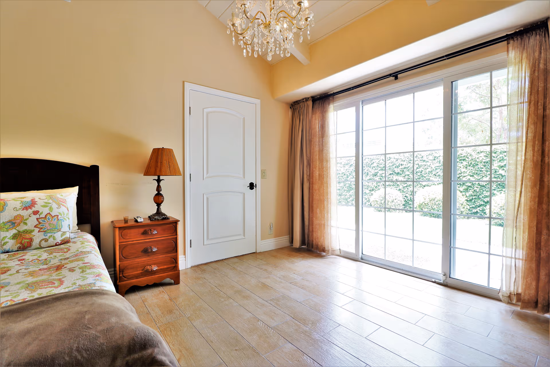 A bright bedroom with a single bed featuring floral patterned bedding, a wooden nightstand with a lamp, a closed white door, and large sliding glass doors with sheer brown curtains that open to an outdoor garden area.