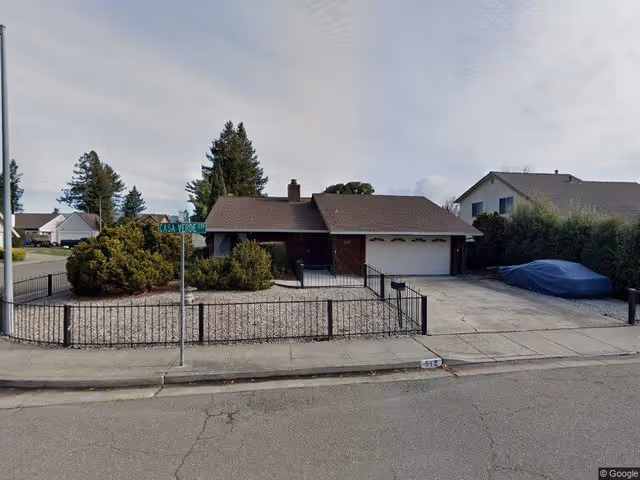 Single-story residential house with a brown roof and attached two-car garage, surrounded by a low black metal fence and gravel landscaping. A street sign labeled Casa Verde Dr is visible in front of the house. A car covered with a blue tarp is parked on the driveway.