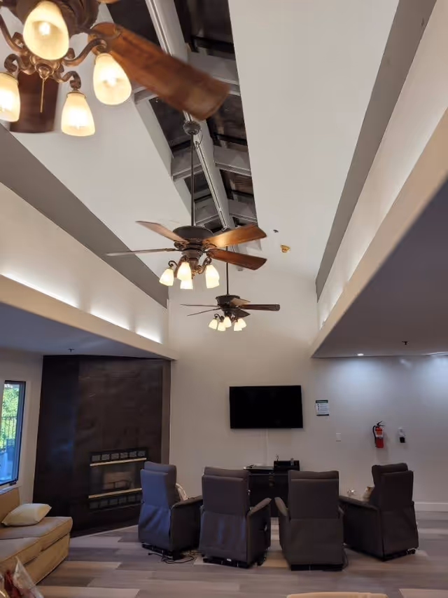 Interior view of a living room area with a high ceiling featuring exposed beams and three ceiling fans with lights. The room has a wall-mounted TV above a small cabinet, four cushioned chairs arranged in a semi-circle facing the TV, a fireplace with a dark surround, and a beige sofa with a pillow near a window.