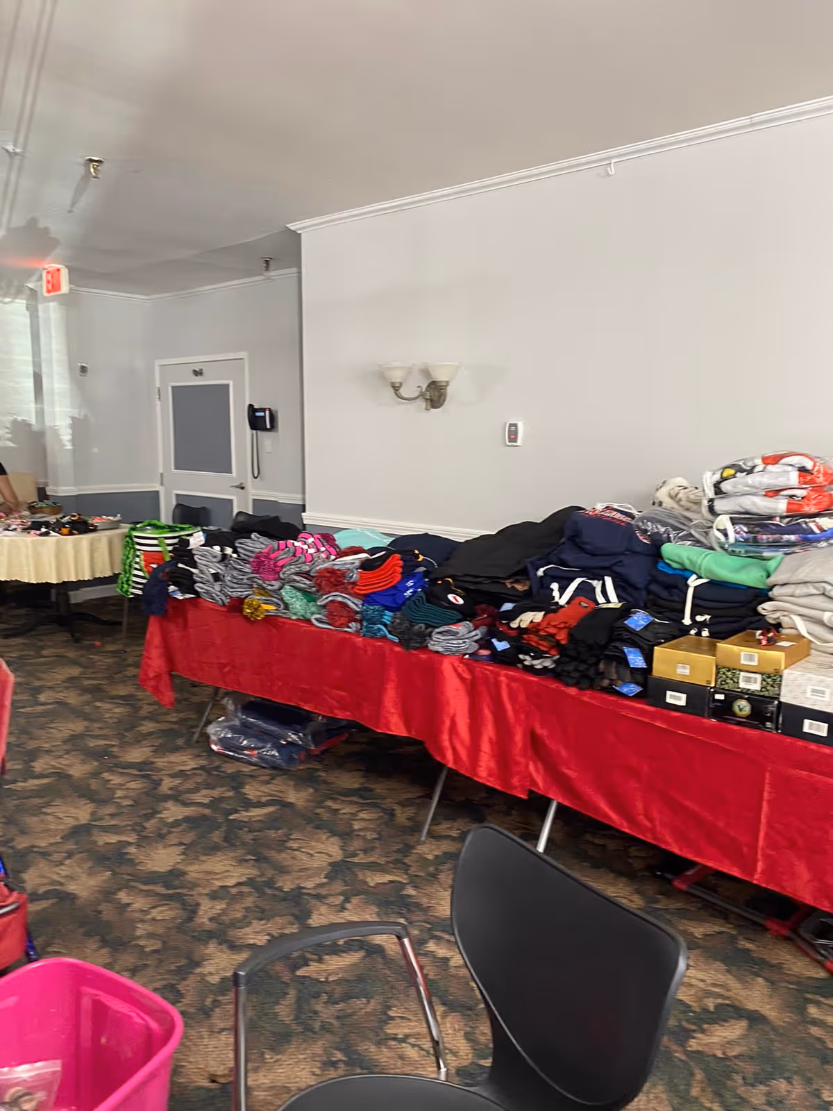 Interior room with tables covered in various folded clothing items and boxes. The tables have red and beige tablecloths. The room has patterned carpet, white walls, a wall-mounted light fixture, and a door with a phone mounted on the wall beside it.
