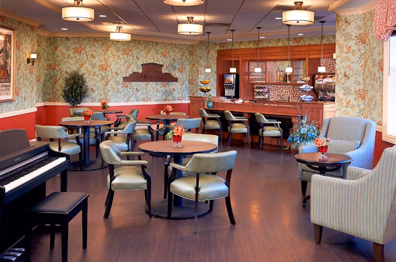 A cozy and well-lit common area with round tables and chairs arranged for seating, a piano with a bench on the left, floral wallpaper with a red lower wall, a counter with bar stools and hanging pendant lights in the background, and a window with patterned curtains on the right.