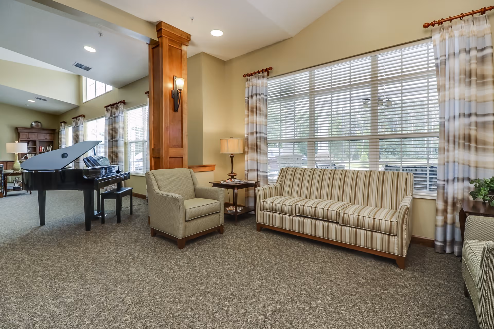 A bright and spacious living room area with a striped sofa, an armchair, a side table with a lamp, large windows with blinds and patterned curtains, and a black grand piano in the background. The room has beige walls, carpeted flooring, and wooden accents.