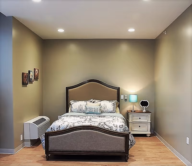 A neatly made bed with a decorative headboard and footboard in a bedroom with beige walls and wooden flooring. There is a nightstand with a blue lamp and a small decorative item on the right side of the bed. Three small framed pictures hang on the left wall, and a heating or air conditioning unit is positioned below them.