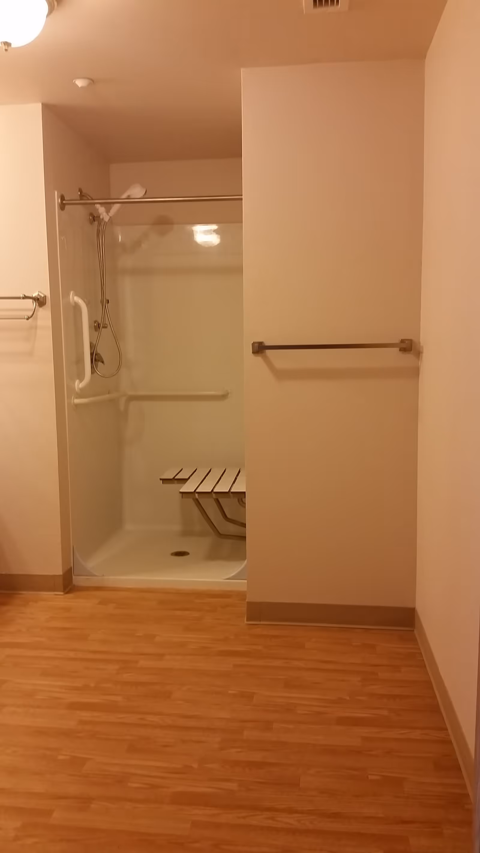 Accessible shower stall with a fold-down bench, grab bars, handheld showerhead, and towel bars in a room with wood-look flooring.