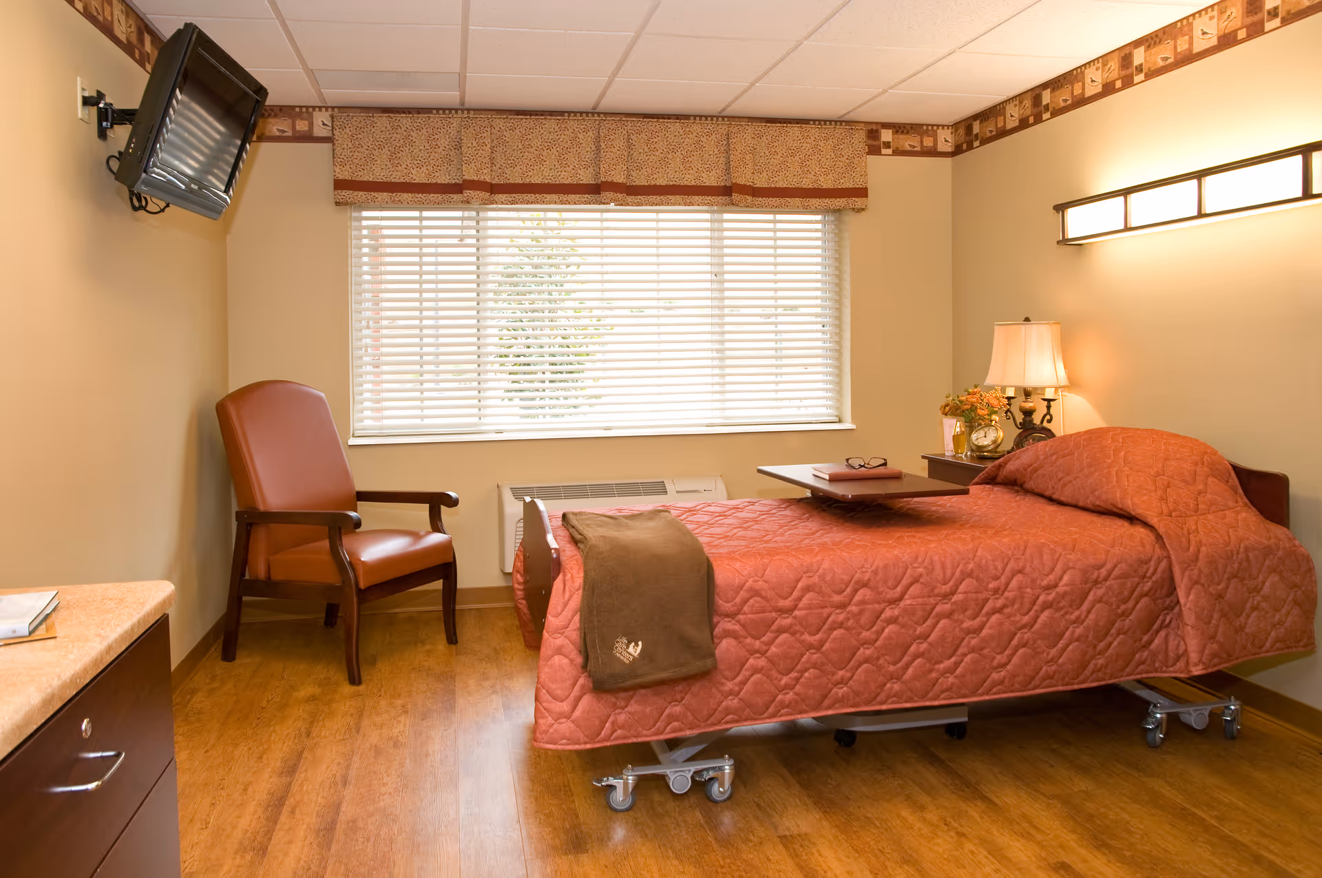 A cozy senior living bedroom with a single bed covered in a rust-colored quilt, a brown blanket folded at the foot, and a small overbed table with a book and glasses. There is a wooden armchair with brown leather upholstery next to a large window with blinds and a patterned valance. A nightstand with a lamp and flowers is beside the bed. A wall-mounted TV is angled above the chair, and the room has warm wooden flooring and beige walls.