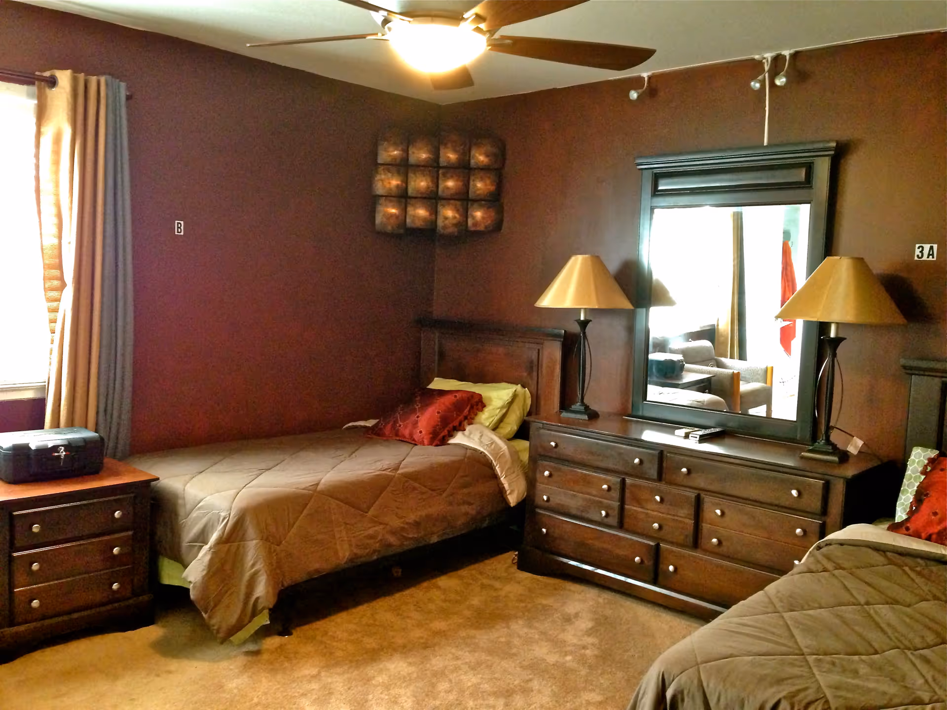A cozy bedroom in an assisted living facility with two single beds, each with brown bedding and decorative pillows. Between the beds is a wooden dresser with a large mirror and two matching lamps. A small wooden nightstand with drawers is next to one bed, and a window with beige curtains lets in natural light. The walls are painted a deep brown color, and a ceiling fan with a light is mounted above.