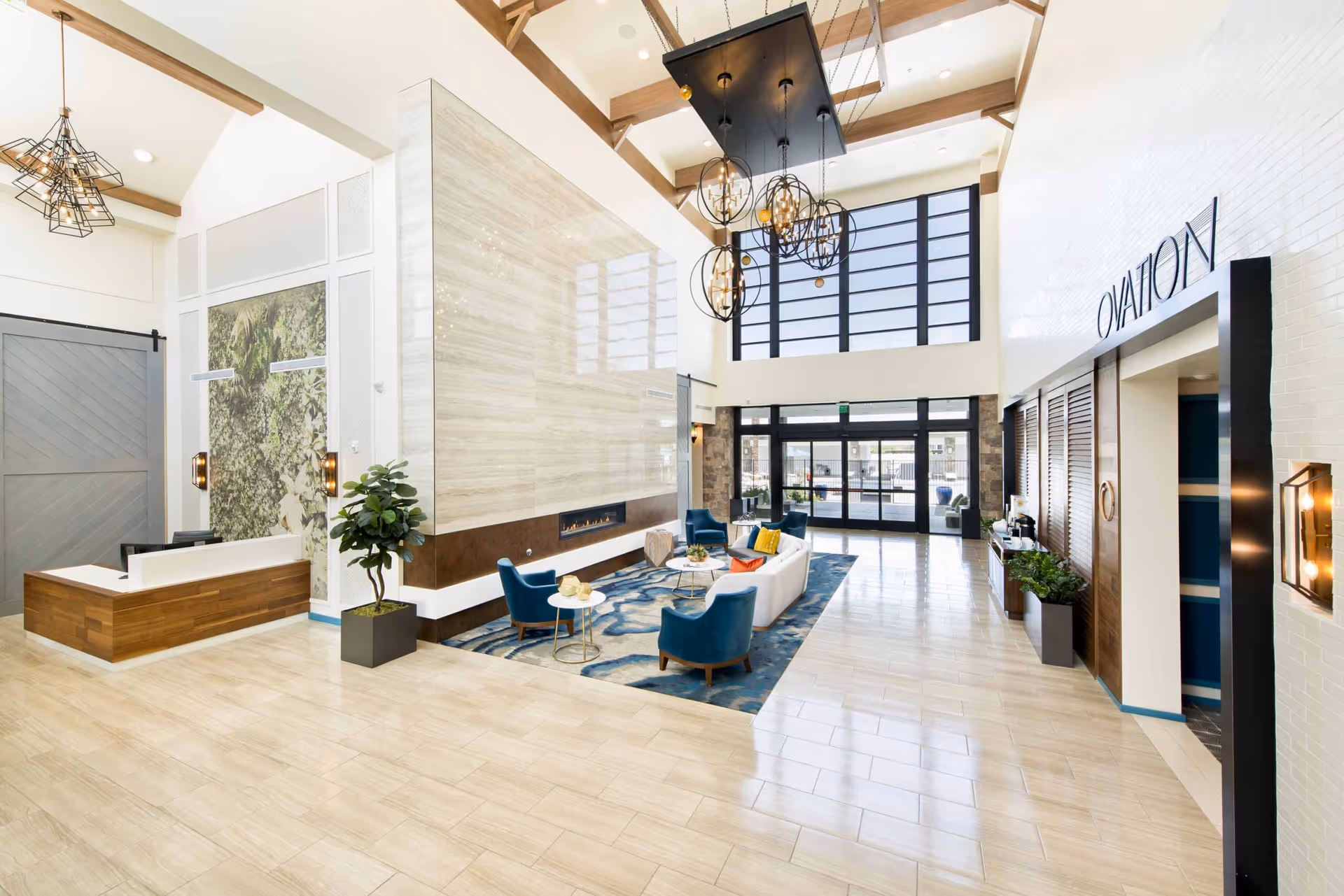Bright and spacious senior living facility lobby with high ceilings, large windows, modern chandeliers, a seating area with blue chairs and a white sofa on a blue patterned rug, a fireplace, a wooden reception desk, and a doorway labeled 'OVATION'.